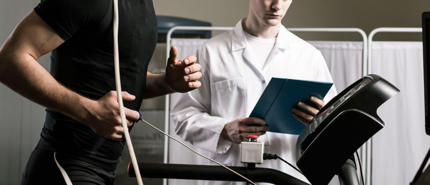 A patient running on a treadmill while a doctor holding a chart observes.